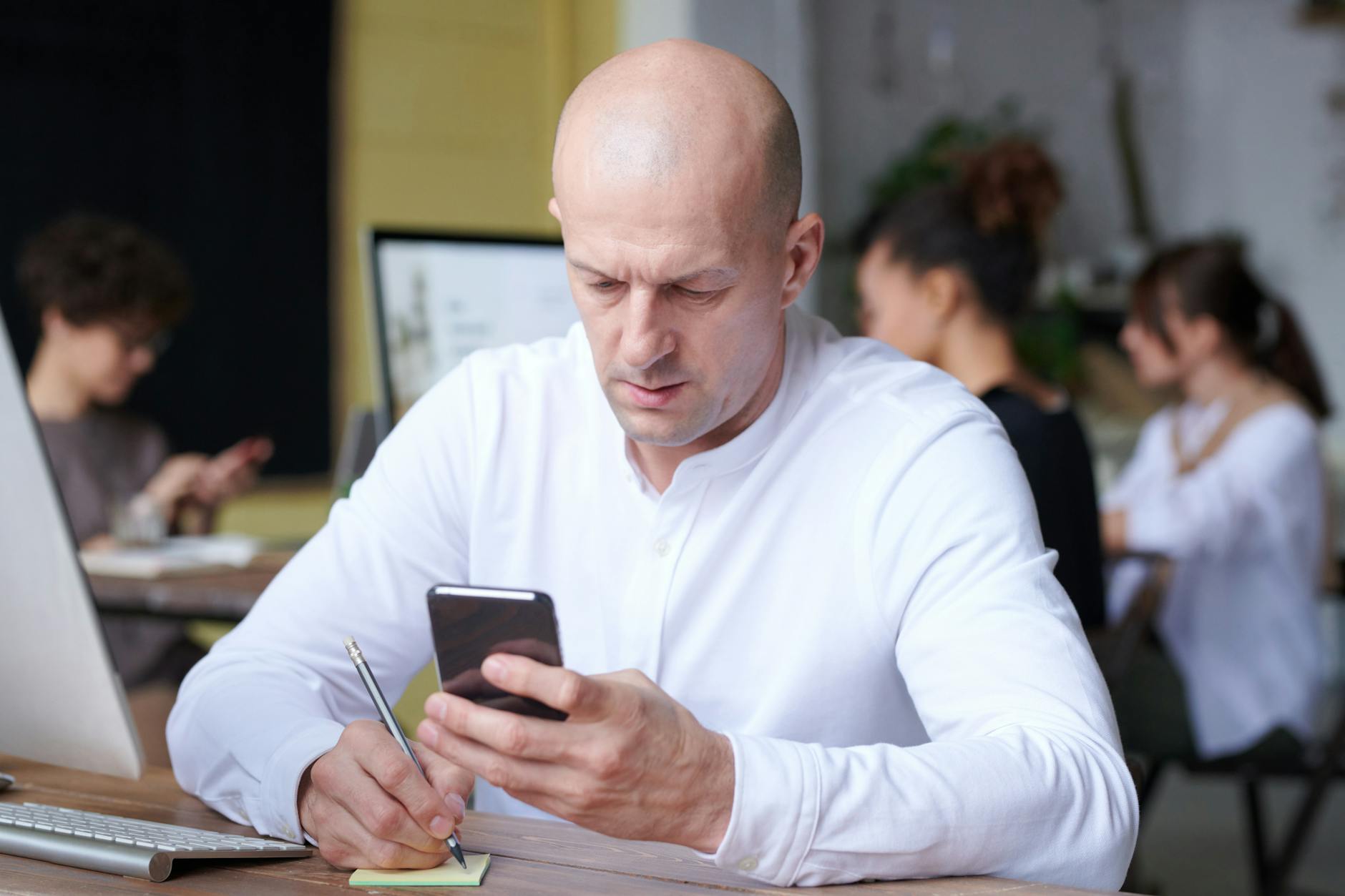 Bald adult in a white shirt writes notes using a smartphone in a busy office setting.
